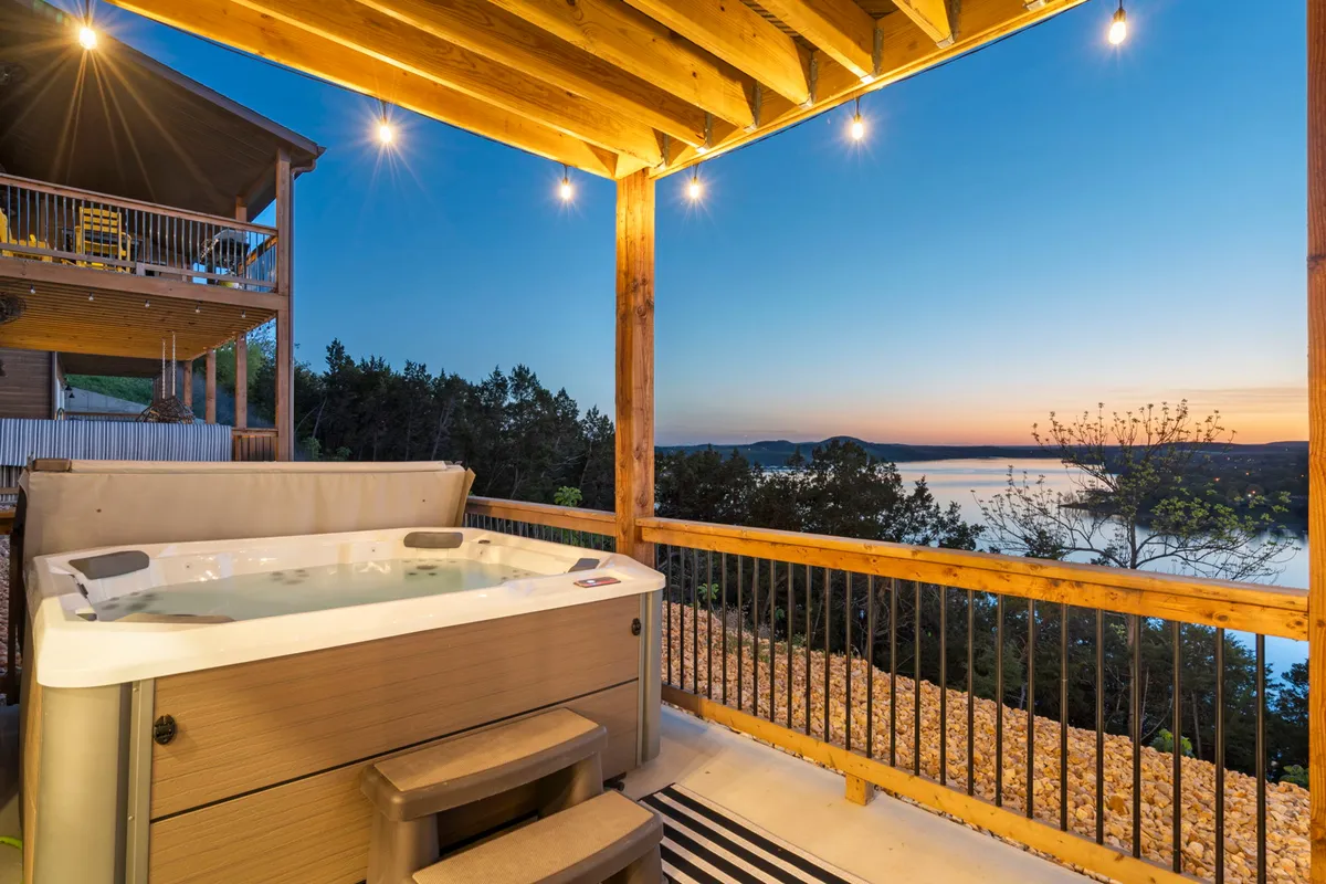 A tranquil hot tub sits on a deck overlooking the lake, adorned with twinkling outdoor lights. The vista extends to the water and forests beyond, creating an inviting spot for relaxation after a day filled with adventure.