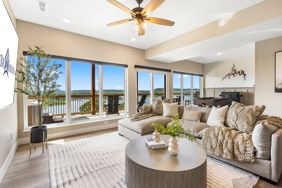 This image features a spacious open-concept living area with a large, comfortable sectional sofa set beside expansive windows that frame panoramic lake views. The stylish coffee table and decorative plant add a designer touch, and the overall mood is cozy and welcoming, inviting conversation or quiet reflection.