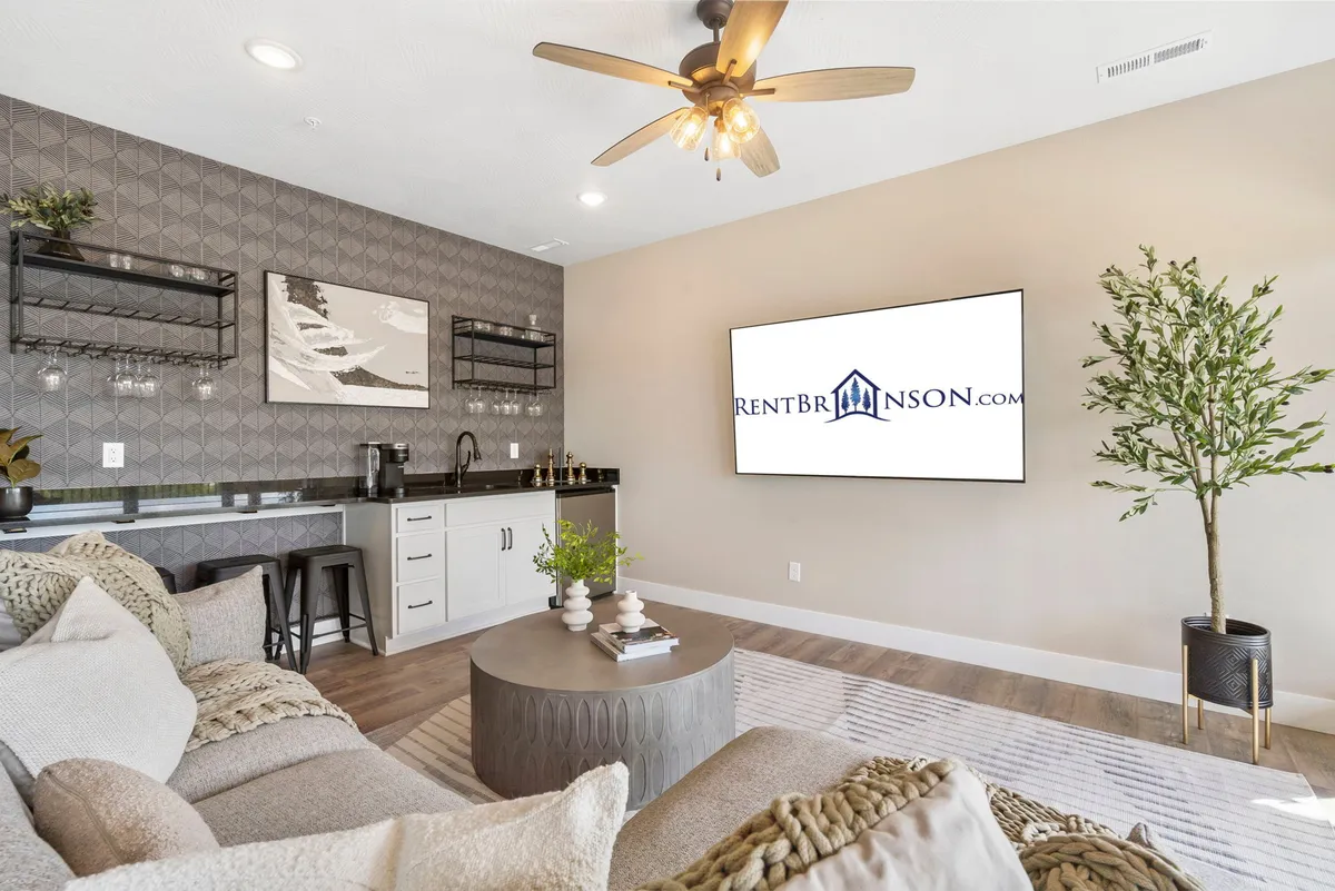 A cozy, contemporary downstairs lounge anchored by a spacious sectional sofa facing a large TV. The modern bar area is visible, complete with shelving, glassware, and decorative plants, conveying an inviting space for socializing, movie nights, and casual gatherings.