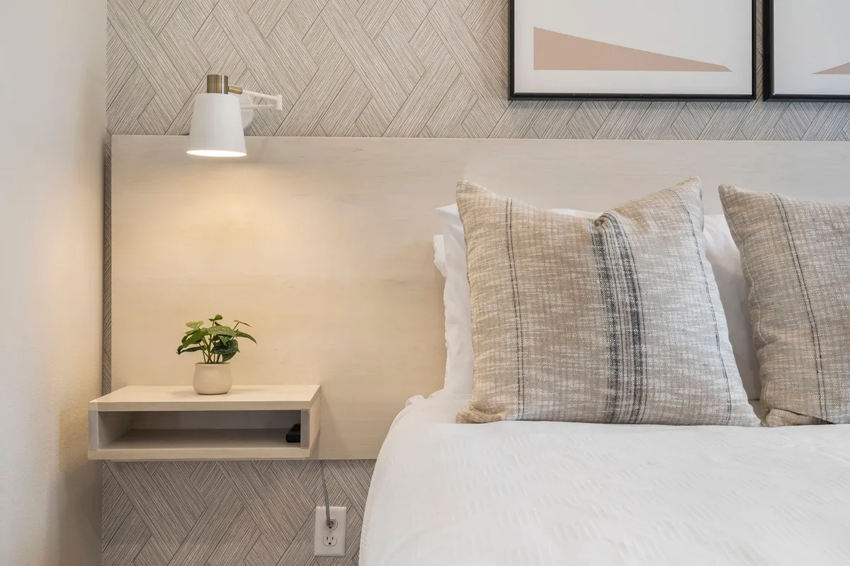 A bright, tranquil bedroom featuring a plush bed with decorative pillows, a nightstand adorned with a potted plant, and wall art above the headboard. Sunlight streams in through the window, illuminating the cozy and inviting space.