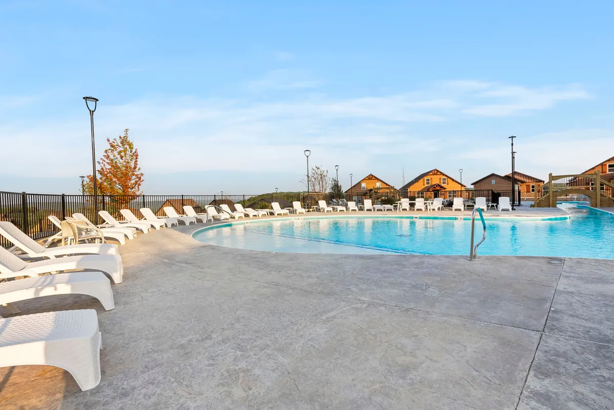 A sun-drenched swimming pool surrounded by comfortable lounge chairs, with cozy cabins and welcoming lamp posts in the background. Guests are seen enjoying the water and relaxing poolside, suggesting a lively yet leisurely atmosphere.