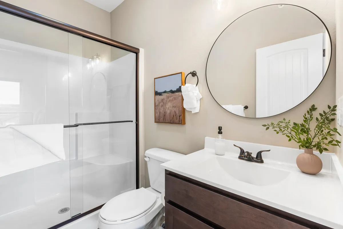 A modern ensuite bathroom featuring a glass shower enclosure, round mirror, vanity with sink, framed artwork, and a plant in a vase. Sunlight brightens the space, evoking a spa-like, serene atmosphere.