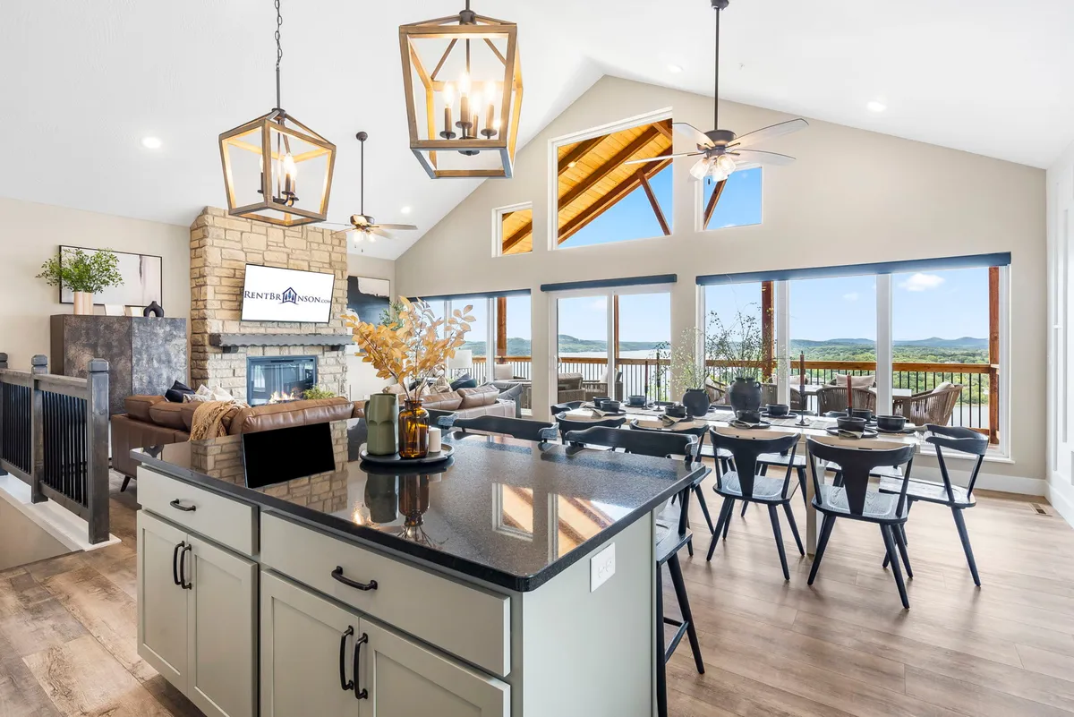 A wide-angle view from an open-plan living and dining space, where large windows provide breathtaking views of the lake. The area features a kitchen island, a dining table, and a stone fireplace, with natural light streaming in, emphasizing the seamless indoor-outdoor flow.