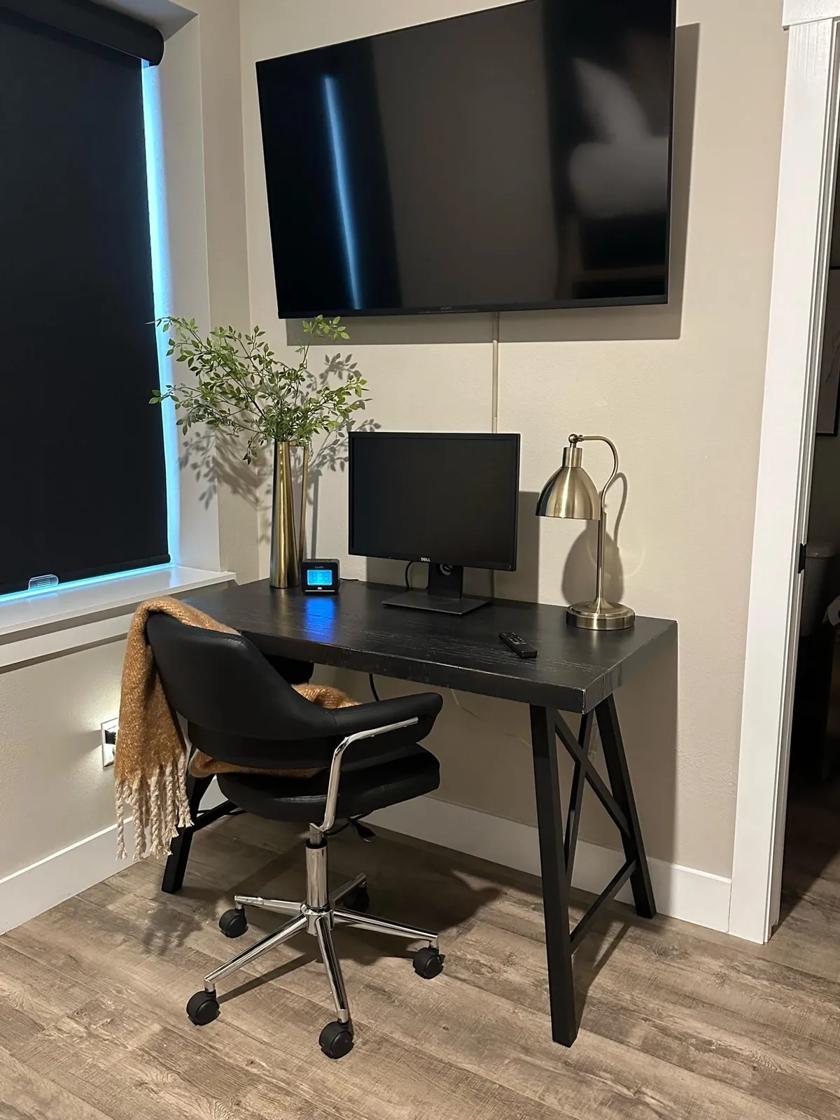 An inviting, well-equipped workspace featuring a desk with a computer monitor, a television, a desk lamp, and a potted plant. The setup is comfortable and thoughtfully arranged for productivity, bathed in soft natural light.