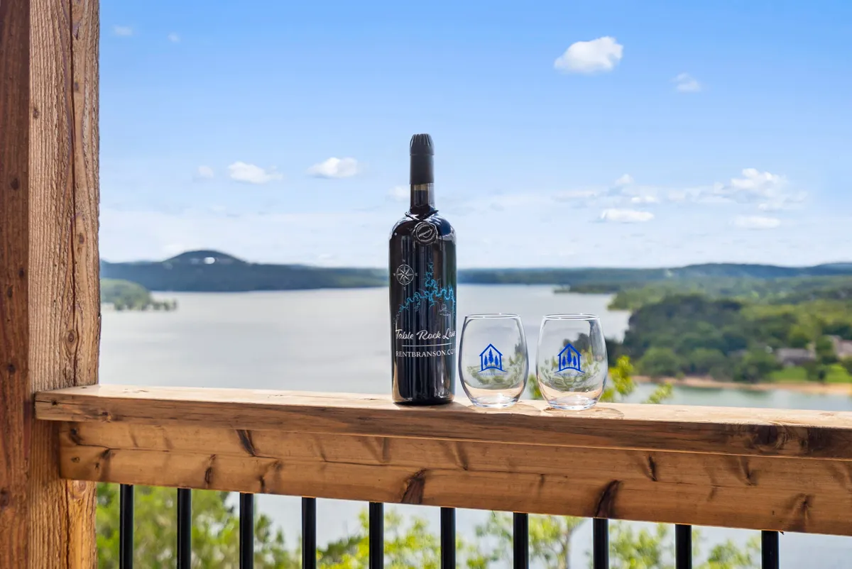 A bottle of wine and two glasses rest on a table, with a stunning scenic lake view in the background. The scene is serene and inviting, suggesting moments of relaxation and celebration with glimpses of the lake even from inside.