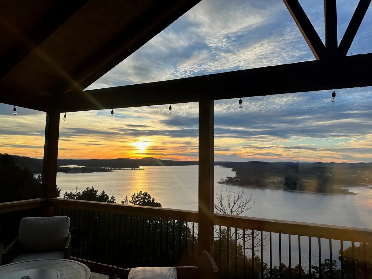 The image showcases a tranquil sunset over Table Rock Lake, framed by rolling mountains in the distance and a spacious wooden patio. The patio invites relaxation and connection, perfect for unwinding with a book or sharing an intimate dinner as the sky glows with evening colors.